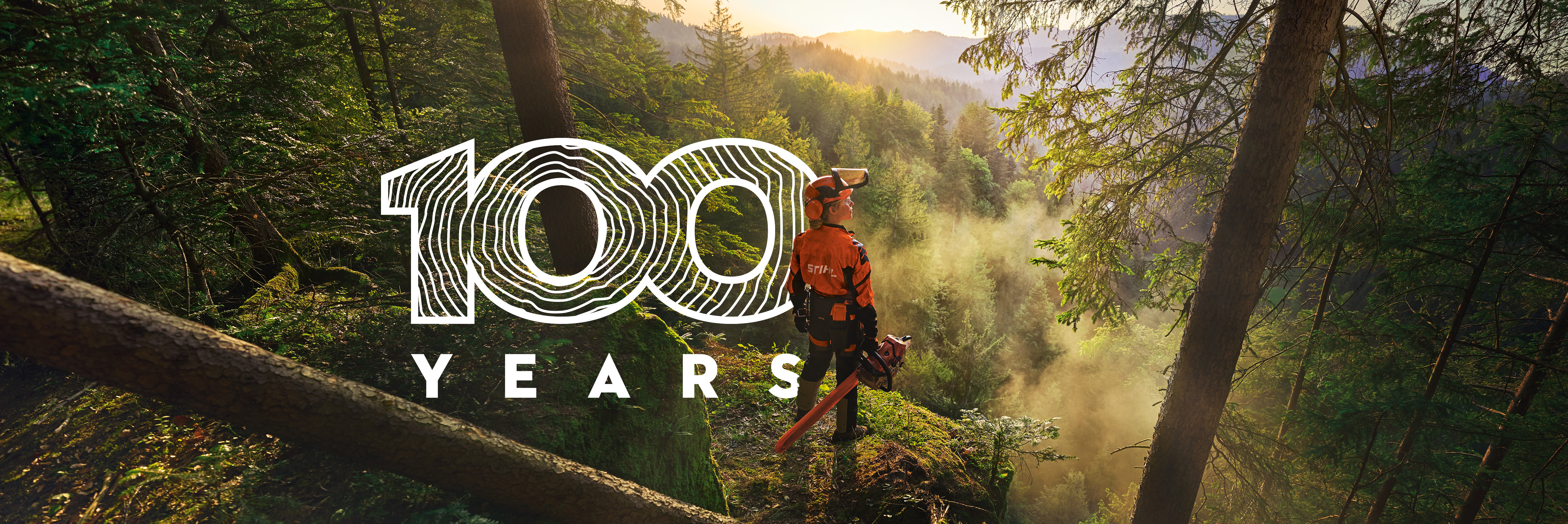 A woman is standing at the edge of a cliff in a saturated green forest. She is wearing orange STIHL workwear, the ADVANCE ProCom Headset, and holding a STIHL MS Chainsaw.
