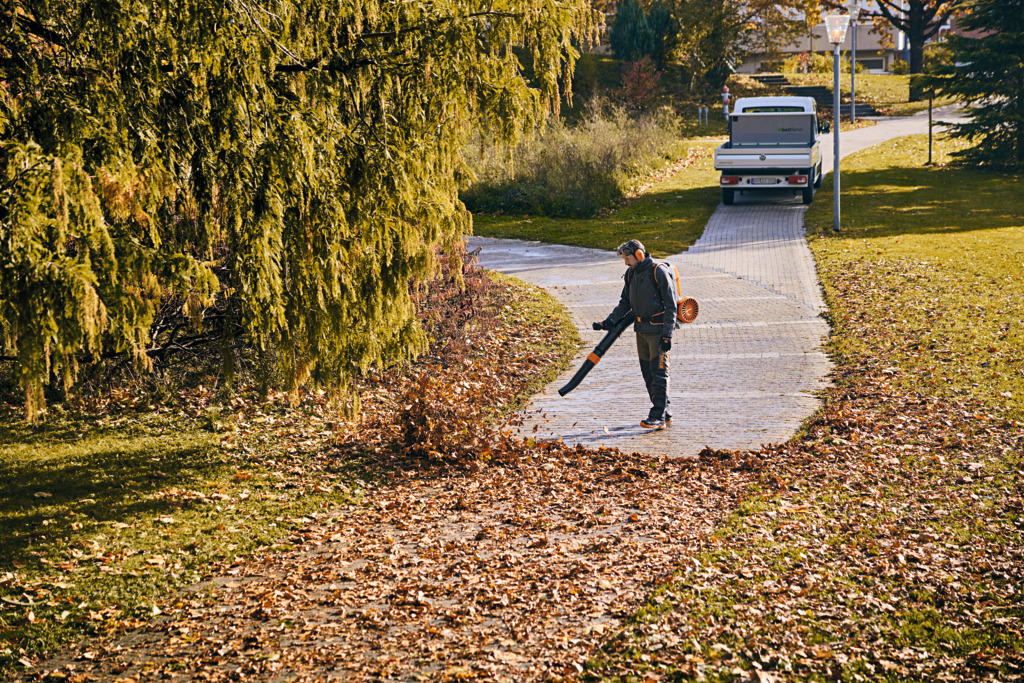 A man in work equipment using a cordless leaf blower to remove leaves from a park path