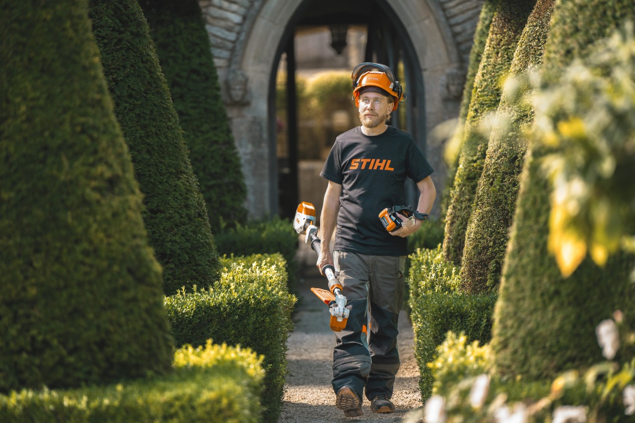 Head gardener Andrew Wain walks through a garden path wearing a safety helmet, a STIHL T-shirt while holding a STIHL battery power tool with a separate battery in his hand