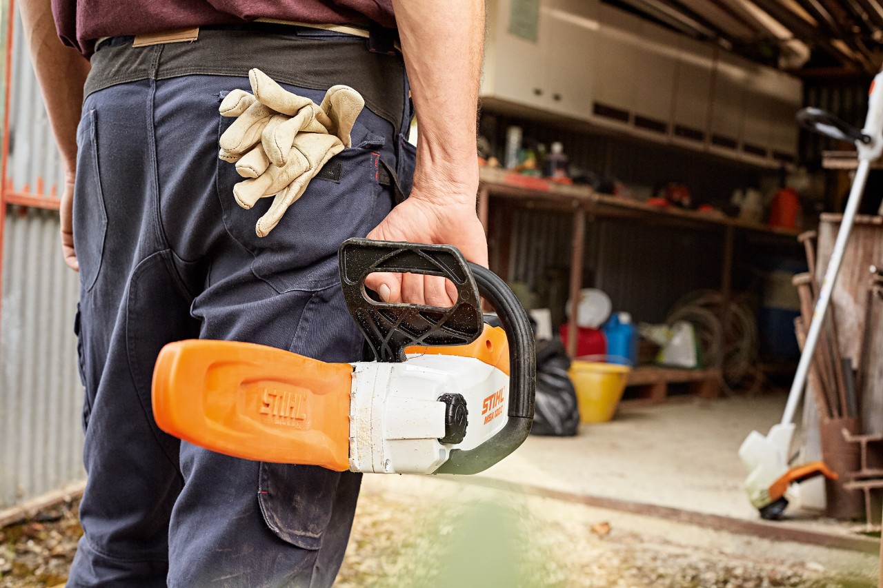 A man walking into a workshop carrying a STIHL cordless chainsaw with scabbard