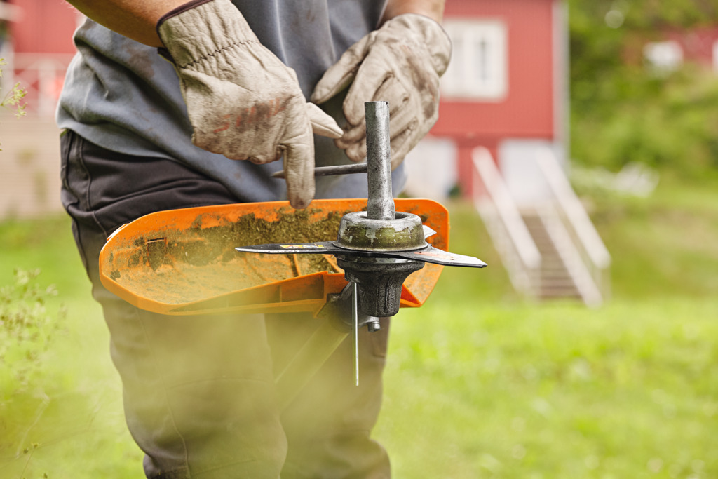 A grass trimmer being held upside down as someone wearing gloves inspects the metal cutting tool